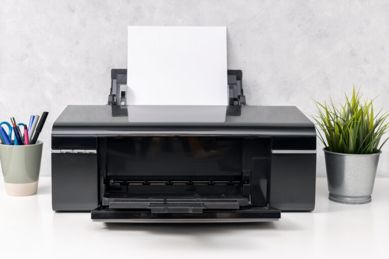Drucker mit weißem Papier. Links eine Tasse mit Stiften, rechts eine Pflange